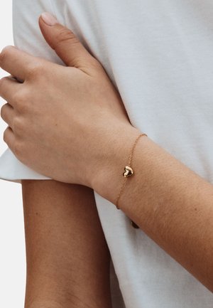 Bracciale con cuore d'oro su un polso, caratterizzato da una catena delicata e una finitura liscia e lucida. La mano poggia su una camicia bianca.