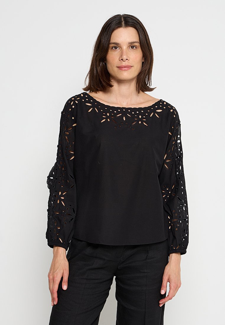 Kobieta z ramionami długości brązowych włosów, nosząca czarną bluzkę z długimi rękawami z kwiatowymi wycięciami na rękawach i dekolcie.