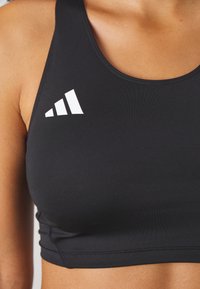 Gros plan d’une personne portant un soutien-gorge de sport noir avec un logo Adidas blanc sur la partie gauche de la poitrine.