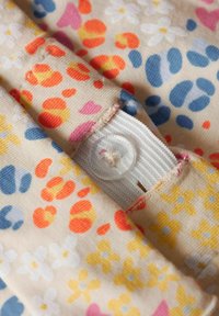 Tkanina w kwiatowy wzór w pastelowych kolorach z pomarańczowymi, niebieskimi i żółtymi kwiatami. Widoczna biała gumka z przezroczystym guzikiem.