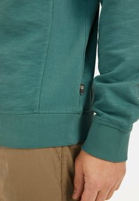 Teal sweatshirt with ribbed cuffs and hem, featuring a side panel textured design and a small multicolor label near the hem.