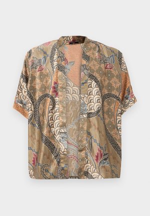 Mann trägt dunkle Jeans, schwarze Schuhe, ein marineblaues T-Shirt, ein gemustertes offenes Hemd und eine schwarze Umhängetasche mit Riemen über der Schulter.