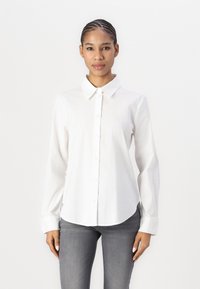 White cotton button-up shirt with a pointed collar, long sleeves, and curved hem, paired with fitted gray jeans.