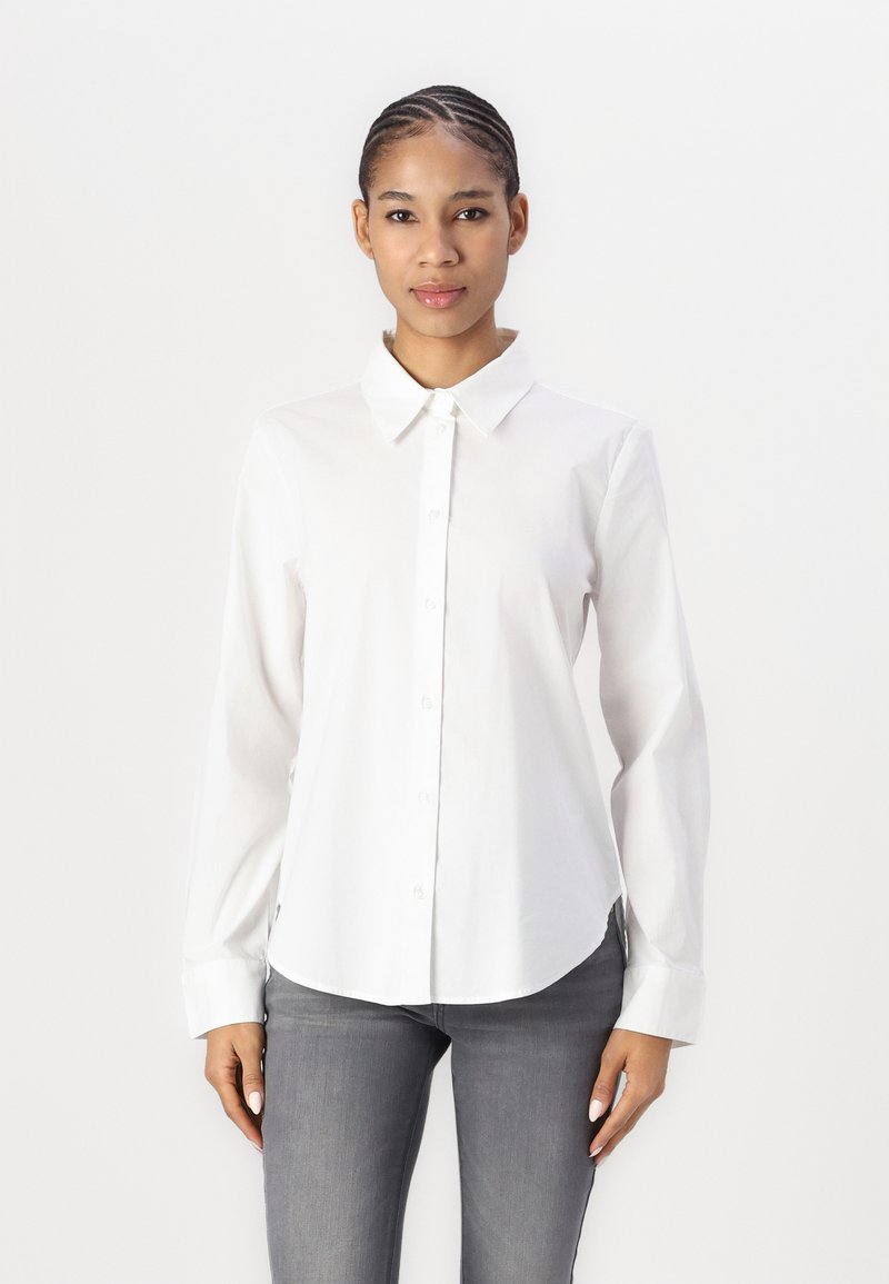 White cotton button-up shirt with a pointed collar, long sleeves, and curved hem, paired with fitted gray jeans.