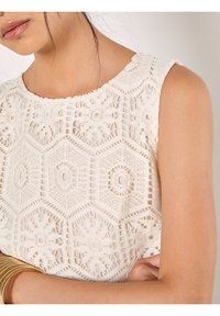 Haut en dentelle crème avec des motifs hexagonaux et des motifs floraux. Doté d'un col rond et d'une finition texturée. Accentué par des bracelets en or.