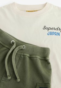 Balta kokvilnas T-kreklā ar uzrakstu "Superdry JAPAN" zilā un melnā krāsā. Olīvzaļas sporta bikses ar auklu un ribotu vidukli.
