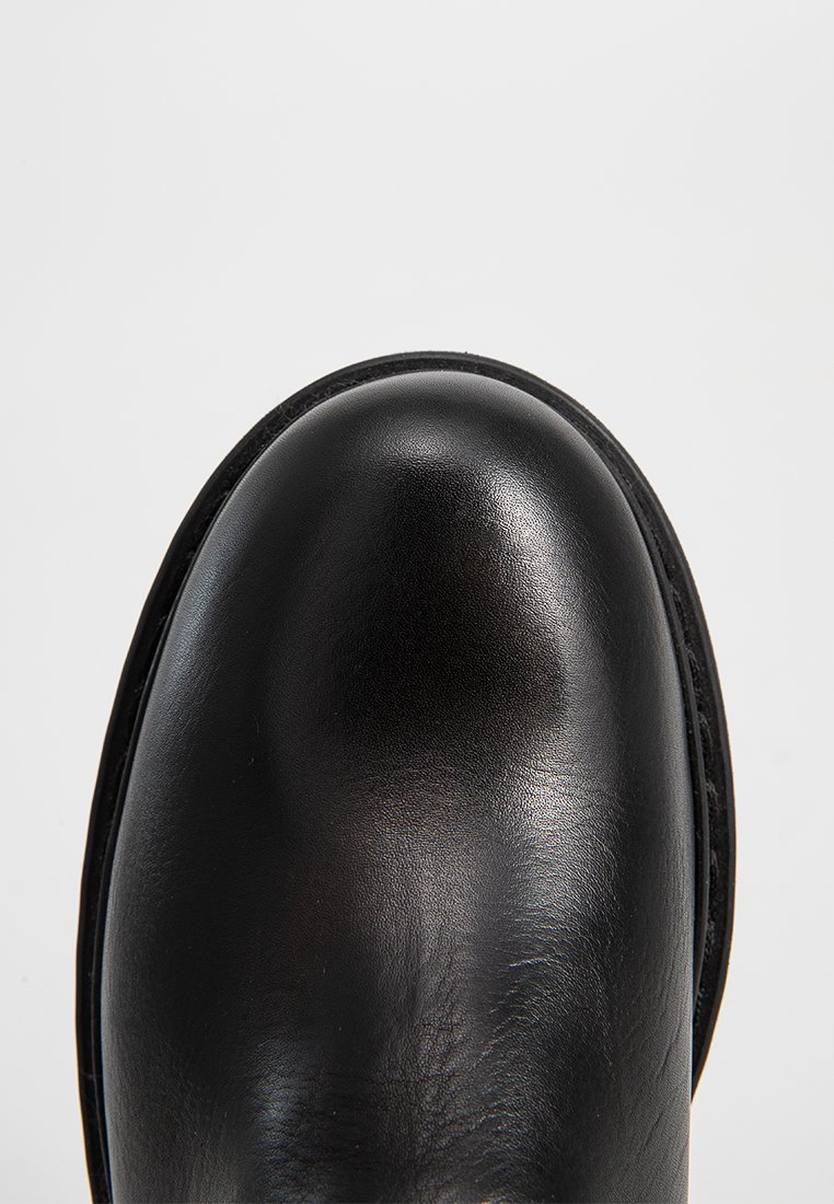 Gros plan sur l'embout d'une botte en cuir noir lisse de forme ronde, avec des coutures visibles le long du bord, sur fond uni.