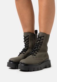 Bottines vert olive avec semelle à crampons noire. Les caractéristiques comprennent un laçage à l'avant, une fermeture éclair sur le côté et des détails de couture contrastants.