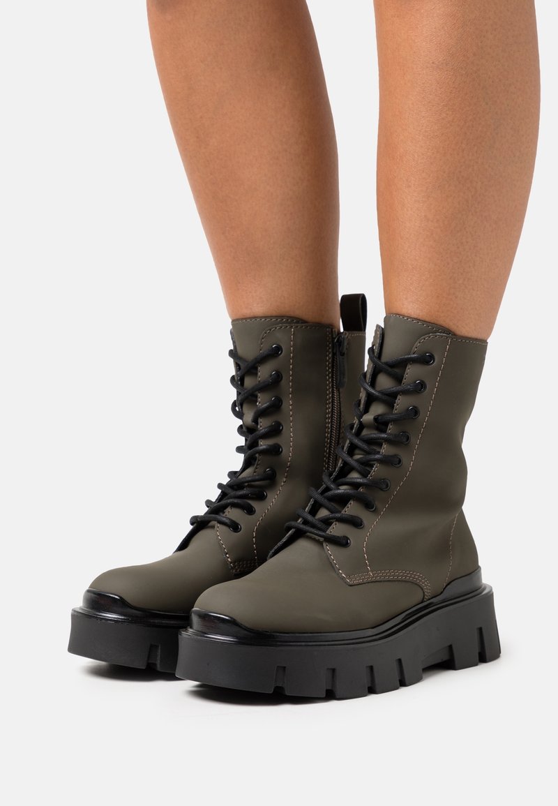 Bottines vert olive avec semelle à crampons noire. Les caractéristiques comprennent un laçage à l'avant, une fermeture éclair sur le côté et des détails de couture contrastants.