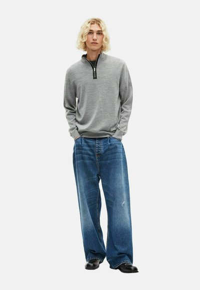 Jongere met schouderlang blond haar, gekleed in een grijze kwartrits trui, losse blauwe jeans en zwarte schoenen, staand met handen in de zakken.