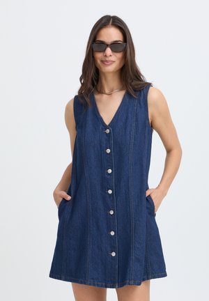Femme portant une robe en jean sans manches à boutons devant, les mains dans les poches et des lunettes de soleil, debout devant un fond clair uni.