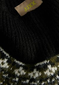 Nahaufnahme von schwarzem Strickstoff mit grün-weißen Mustern aus Wolle und einem beigen JJXX-Markenlabel, das auf das Kleidungsstück genäht ist.