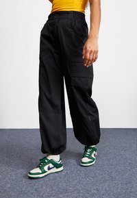 Svarta cargobyxor med elastiska muddar, med flera fickor och en avslappnad passform. Kombineras med gröna och vita sneakers.