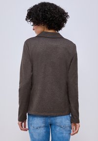 Personne aux cheveux bouclés et foncés, portant un haut à manches longues marron foncé et un jean bleu clair, vue de dos sur un fond clair uni.