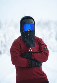 Czerwona kurtka narciarska z czarnymi akcentami i kapturem, z logo na piersi, w połączeniu z czarnymi rękawiczkami i niebiesko zabarwionymi goglami narciarskimi.