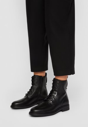 Schwarze Leder-Stiefeletten mit Schnürung und seitlichen Reißverschlüssen, getragen zu schwarzen, gekürzten Hosen auf hellem grauem Hintergrund.