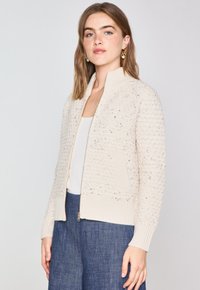 Cardigan en tricot couleur crème avec un motif texturé, des bords côtelés et des poches latérales. Présente une fermeture éclair à l'avant et un col montant.