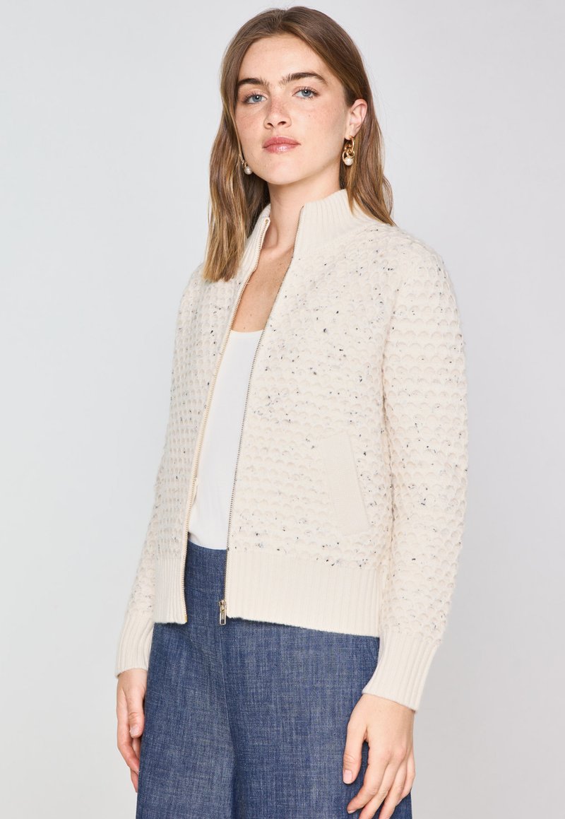Cardigan en tricot couleur crème avec un motif texturé, des bords côtelés et des poches latérales. Présente une fermeture éclair à l'avant et un col montant.