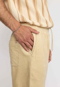 Beige corduroy broek met een ribbelstructuur, elastische tailleband en een zijzak met rits. Vergezeld van een gestreepte beige en oranje top.