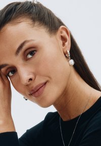 Sudraba riņķveida auss gredzeni ar pērļu akcentu, ar pulētu apdari. Modelis valkā melnu blūzi, izceļot auss gredzenus uz gaiša fona.