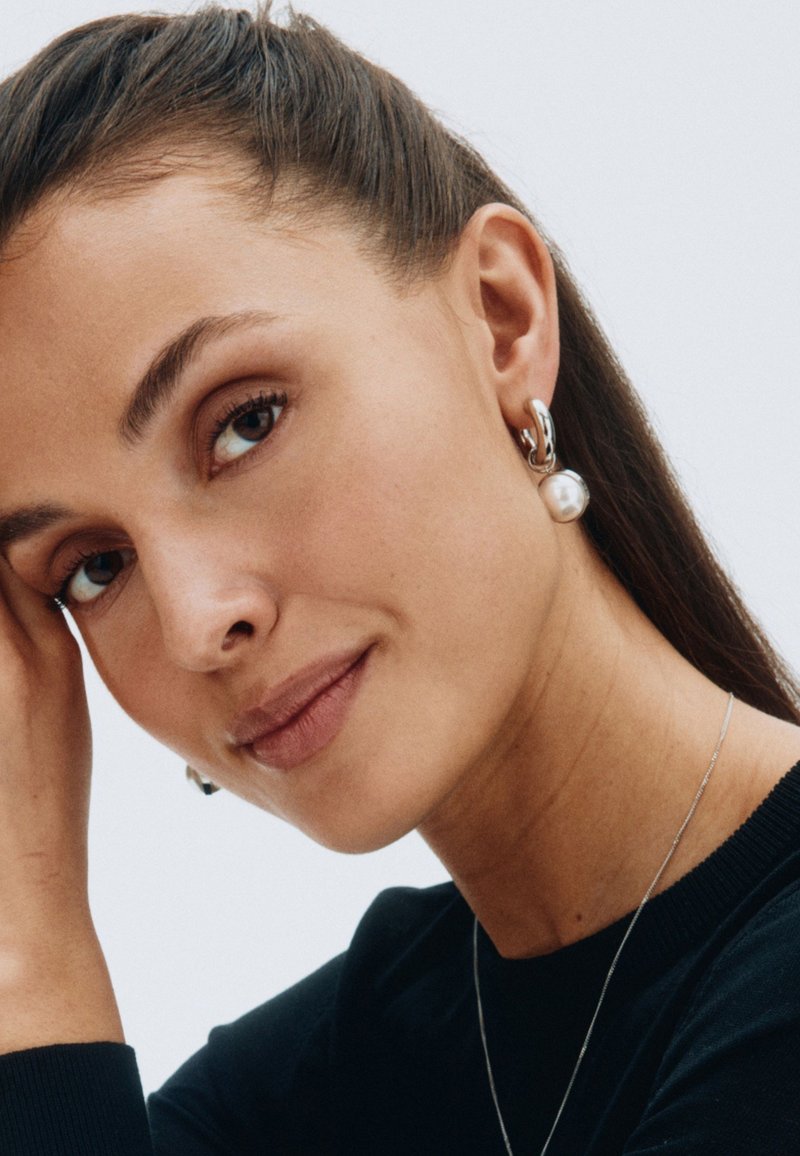 Sudraba riņķveida auss gredzeni ar pērļu akcentu, ar pulētu apdari. Modelis valkā melnu blūzi, izceļot auss gredzenus uz gaiša fona.