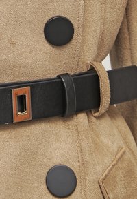 Karmelowy zamszowy płaszcz z czarnymi guzikami i szerokim czarnym paskiem z prostokątną złotą klamrą. Gładka textura z detalicznym przeszyciem.