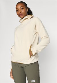 Crème fleece pullover met een hoge kraag, bruine afwerking en kangoeroe zakken; gecombineerd met olijfgroene broek met een logo op het been.