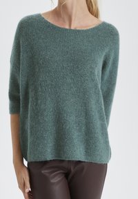 Light teal, oversized knit sweater with a wide neckline, three-quarter sleeves, and a soft, textured finish. Paired with brown leather pants.