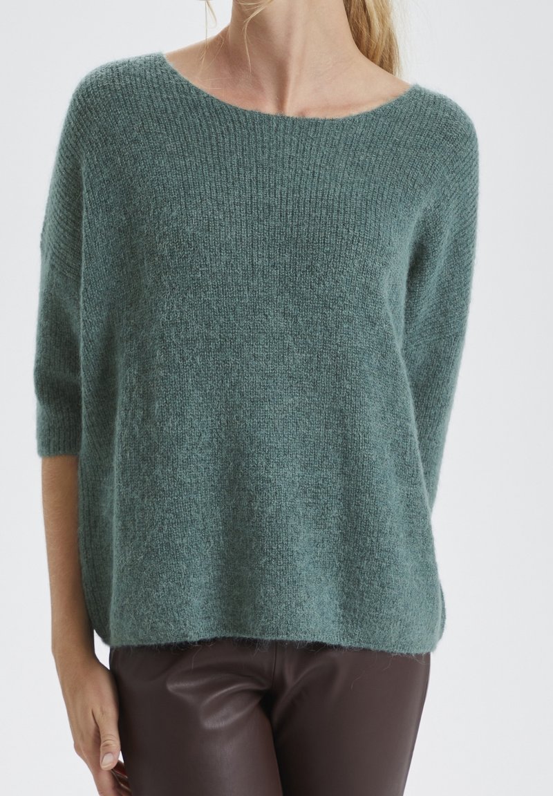 Light teal, oversized knit sweater with a wide neckline, three-quarter sleeves, and a soft, textured finish. Paired with brown leather pants.
