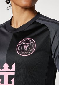 Black soccer jersey featuring a pink club logo and crown emblem, with a sporty design and textured fabric. Three gray stripes along the shoulders.