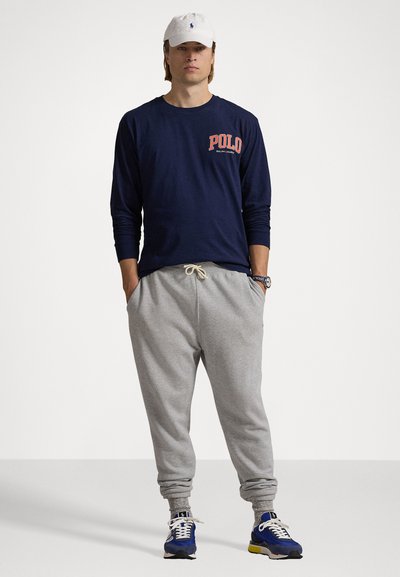 Jūrinis ilgarankovis marškiniai su raudonu "POLO" tekstu, pilkos sportinės kelnės ir mėlyni sportiniai batai. Balta kepurė ir laikrodis ant riešo. Kasdienis apranga.