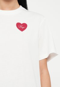 Camiseta blanca de manga corta con un pequeño corazón rojo y la palabra "Hugo" en cursiva en la parte superior izquierda del pecho.