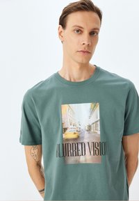 Camiseta de algodón verde con un gráfico central que presenta una escena callejera difusa y el texto "VISIÓN DIFUSA" en letras negras en negrita.
