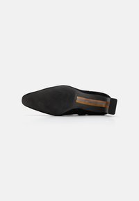 Zapato de tacón negro con una textura suave y punta afilada. Presenta una suela negra sólida y un detalle de rayas marrones cerca del tacón.