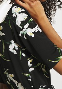 Czarna sukienka w kwiaty z białymi wzorami lilii i zielonymi liśćmi, wykonana z gładkiego materiału, o luźnym kroju i krótkich rękawach.