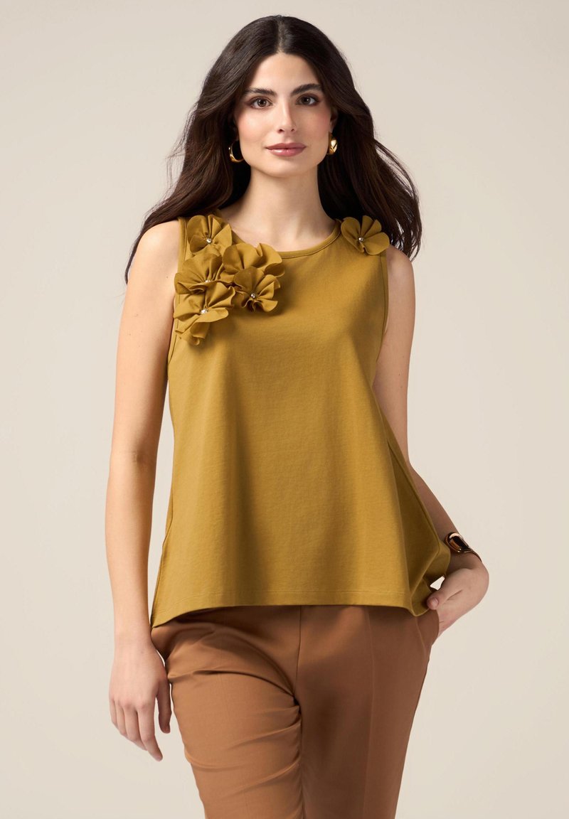 Top senza maniche color senape con una vestibilità ampia, decorato da tre fiori di tessuto sulla spalla sinistra, con accenti di perle. Indossato con pantaloni marroni.