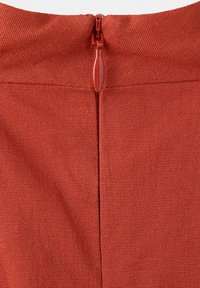 Tissu rouge orangé avec une fermeture éclair fermée et une languette en tissu centrée dans une couture sur un fond uni.