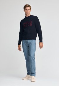 Marinblå stickad tröja med texten "POLO CLUB" i rött, kombinerad med ljusblå denimjeans och beige sneakers, stående mot en enkel bakgrund.