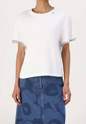 Mujer con una camiseta blanca de manga corta con ribete de pedrería en las mangas y una falda vaquera azul con grandes patrones florales.