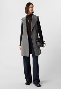 Langer grauer Weste mit Reverskragen, die über einem schwarzen Rollkragenpullover getragen wird. Sie hat eine taillierte Passform und wird mit navyblauen, weiten Hosen und einer schwarzen Tasche kombiniert.