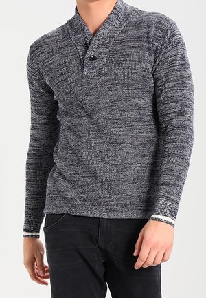 Uomo che indossa un maglione henley grigio mélange a maniche lunghe con bottoni neri e pantaloni neri, in piedi con le mani rilassate.