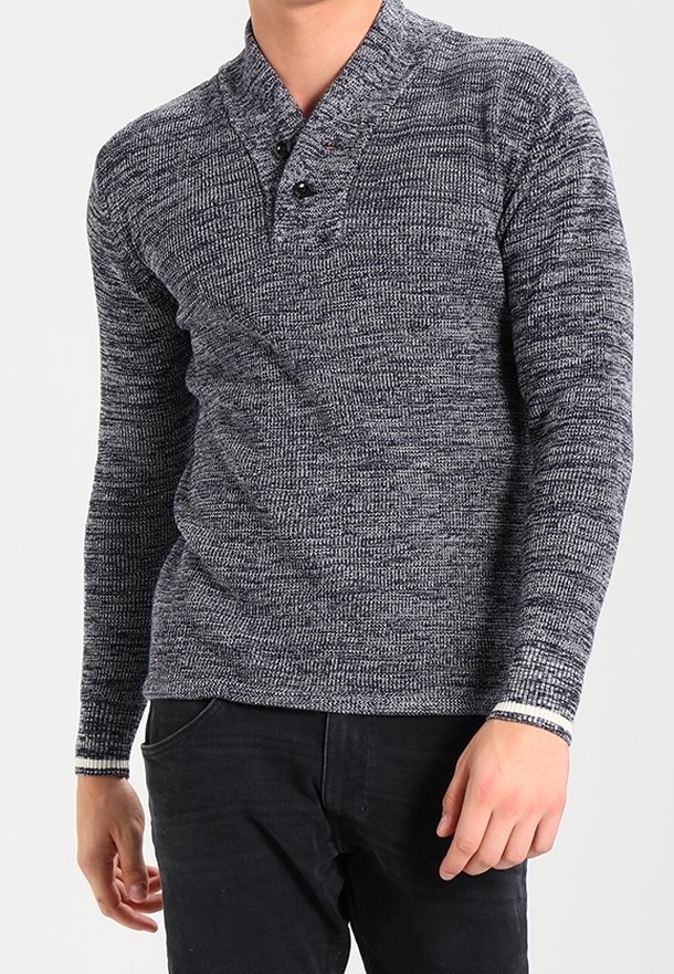 Homme portant un pull henley en maille gris chiné à manches longues avec des boutons noirs et un pantalon noir, debout, les mains détendues.