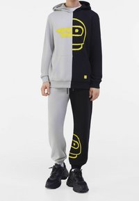 Man med en hoodie i delade färger och sweatpants, grått på vänster sida och svart på höger sida, med gula grafiska mönster och svarta sneakers.