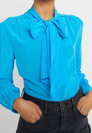 Person wearing a bright blue blouse with a large bow tie neck, long puffed sleeves, and black high-waisted jeans with hands in pockets.