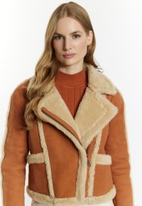 Bruine shearling jas met een fluffy beige kraag, ritssluiting en gestructureerde panels. Draagt over een geribbelde oranje top. Glad suède oppervlak.