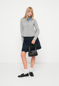 Sivasti rebrast cardigan oblečen preko modre srajce z gumbi, v kombinaciji z zeleno in mornarsko karirasto krilo. Drži črno torbico z teksturo in studenci.