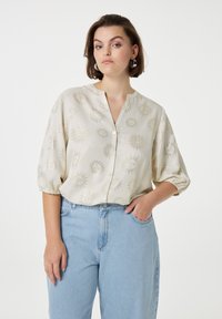 Cremefarbige Bluse mit metallischen Sonnenmustern, Rundhalsausschnitt und Knopfverschluss. Verfügt über geraffte Ärmel und wird mit hellblauen Jeans kombiniert.