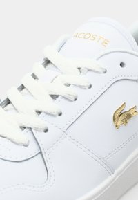 Baltos sportinės avalynės su odiniu viršumi, plokščiomis baltomis raištelėmis, auksiniu Lacoste logotipu ir tekstūruota padanga. Minimalistinis dizainas ir švarus estetikos pojūtis.