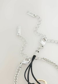 Sølv lænker med en sort flettet snor, udstyret med en løvemedlås og dekorative accenter, herunder rektangulære metalplader med sten.