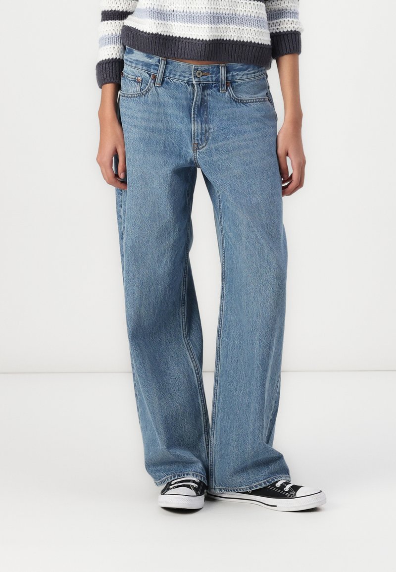 Vaqueros de pierna ancha en un azul claro, hechos de denim, con un diseño clásico de cinco bolsillos y un ligero desteñido. Combinados con zapatillas en negro y blanco.